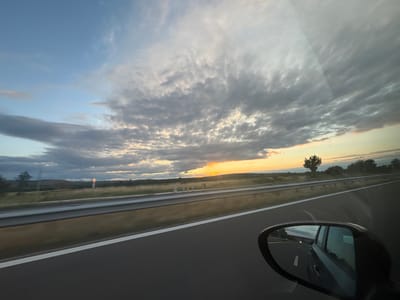 The image depicts a view from a car driving on a highway, likely in or near Kalugerovo, Bulgaria, as suggested by the prompt. The perspective is from the right side of the vehicle, with the side mirror prominently featured in the lower right corner, reflecting the sky and the road behind. The highway itself has a grey asphalt surface with a white lane marker visible. A silver metal guardrail runs along the left side of the highway, separating it from a grassy field. The field is dotted with sparse vegetation and a few trees in the distance. The sky is a significant element, filled with dramatic clouds that are a mix of grey and white, with patches of golden light from the setting sun breaking through. The horizon is marked by a low, undulating landscape. There are no people or animals visible in the scene. The overall impression is one of travel during a picturesque sunset.