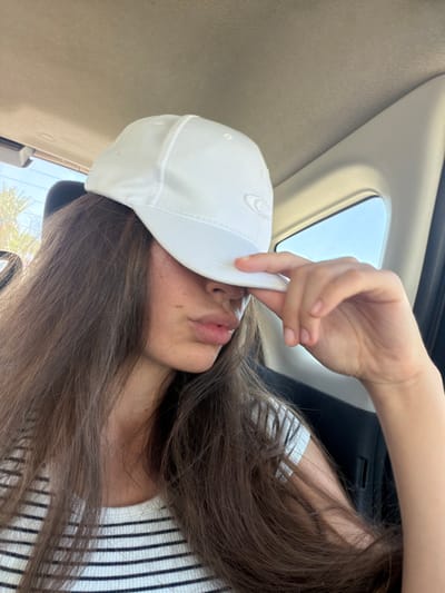 In Larnaca, Cyprus, a young woman with long brown hair is captured in a medium shot, wearing a white baseball cap and a black and white striped shirt. She is sitting in a vehicle, as indicated by the car's interior visible in the background. The woman is holding the brim of her cap with her right hand, obscuring her eyes and nose, and her lips are slightly pursed. The lighting suggests it is daytime, with natural light coming from a window on the right side of the frame. The environment appears to be a typical car interior with fabric and plastic surfaces.