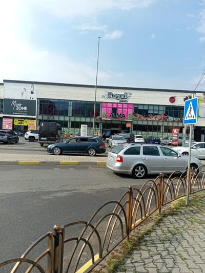 This media file captures a street view in Tiachiv, Ukraine, on a sunny day with a bright blue sky and scattered clouds. The scene is dominated by a large shopping complex with the prominent signage "Royal plaza." The building features a modern facade with large glass windows, reflecting the surroundings.
Several cars are parked and driving on the asphalt road in front of the shopping center. A dark blue station wagon is in the foreground, with a silver station wagon behind it. Other vehicles, including a black van, are also visible. A pedestrian crossing sign stands near the right edge of the frame.
The shopping complex has various advertisements and storefronts. Visible text includes "Brand House Kids," "Men Zone" with a slogan about men's clothing, and "ALMA" with what appears to be a supermarket designation. Other signs advertise "SECRET" products, including perfume, cosmetics, and shoes, as well as "FISH" and "SUSHI." The "VOLT" brand is also displayed.
The street is lined with a decorative metal fence and a sidewalk paved with interlocking stones, with patches of grass growing through. The time of day appears to be daytime, given the bright sunlight and shadows. The overall atmosphere is that of a typical commercial area on a pleasant day.