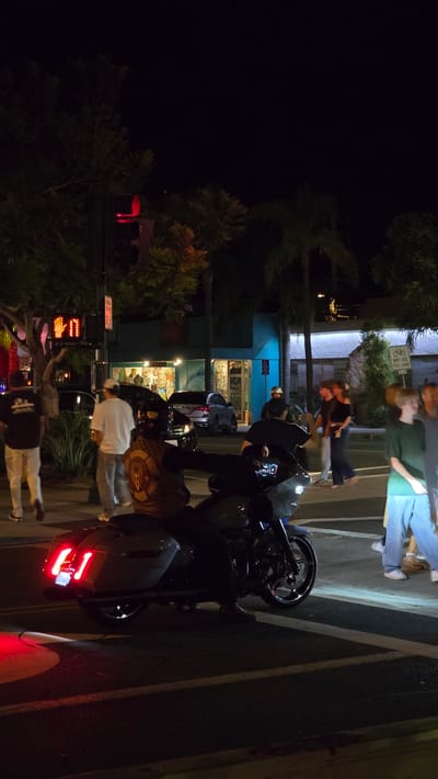 At night in Encinitas, California, a motorcyclist is stopped at a crosswalk. The motorcycle, a white touring model, has its brake lights illuminated and is positioned in the right lane of traffic. The motorcyclist, wearing a dark helmet and a patterned vest, is seated on the vehicle. Several pedestrians are visible on the sidewalk to the left, including a man in a black t-shirt and light pants, and another in a white t-shirt and grey pants. To the right, other pedestrians are walking across the street. In the background, businesses with illuminated storefronts are visible, and palm trees line the street. A traffic signal with a red light is visible overhead.