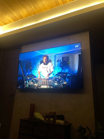 The image shows a television mounted on a wall, displaying a DJ performing. The environment appears to be indoors, possibly a living room, with wood paneling on one side and a shelf with various objects on the other. The exact location in Huixquilucan de Degollado, Mexico cannot be determined without further information.