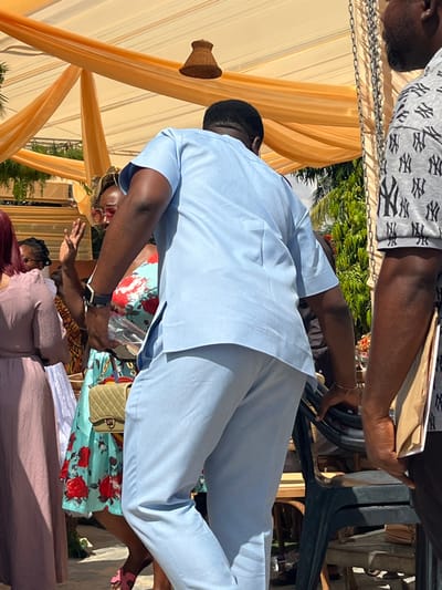 The scene depicts an outdoor social gathering in Kumasi, Ghana, under a draped canopy of light orange fabric. Bright daylight conditions are present. A male individual, positioned centrally with his back partially to the viewer, wears a light blue short-sleeved tunic and matching trousers. A dark smartwatch is visible on his left wrist. Partially obscured behind him, a female individual with red-framed glasses wears a light-colored dress with a red floral pattern; her right hand is raised in a clear gesture. This woman carries a light gold quilted handbag with a chain strap. To the far left, another female is partially visible in a light pink dress. On the right side, a male individual, partially visible, wears a grey t-shirt patterned with repeating white "NY" logos and holds a brown paper item. In the background, green foliage is visible, alongside stacked dark green plastic chairs and other event seating arrangements. A woven basket-like light fixture hangs from the canopy.