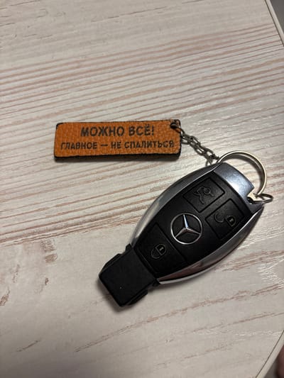 The image displays a Mercedes-Benz car key with a leather keychain attached. The keychain has Russian text embossed on it that reads "МОЖНО ВСЁ — ГЛАВНОЕ — НЕ СПАТЬСЯ", which translates to "Everything is possible - the main thing is not to sleep". The key is black and silver with the Mercedes-Benz logo prominently featured. It has multiple buttons, including what appear to be lock, unlock, and trunk release icons. The scene is set on a light-colored, textured surface resembling wood grain, possibly a table or countertop. The lighting suggests an indoor environment, with no indication of specific time of day or weather conditions. The overall mood is neutral, focusing on the objects themselves. The image is taken from a top-down perspective. The location is Kemerovo, Russia.