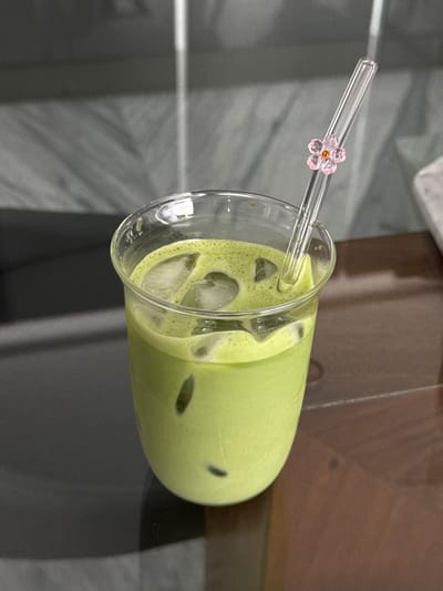 The image displays a transparent drinking glass containing an iced green beverage, consistent in appearance with matcha. Multiple irregular ice cubes are visible within the liquid, and a layer of foam rests on the surface. A clear glass straw, adorned with a small pink five-petaled flower charm featuring an orange center, is inserted into the drink. The glass rests upon a dark brown, reflective surface. The background consists of blurred grey tones with subtle vertical and horizontal linear patterns, suggestive of a textured wall or paneling. This scene is situated in Zapopan, Mexico.