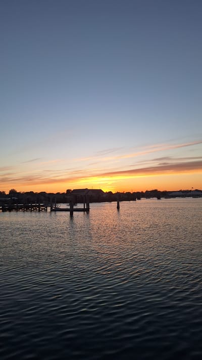 The image depicts a serene waterfront scene at dusk on Nantucket, Massachusetts, United States of America. The sky transitions from a pale blue at the top to a vibrant spectrum of orange and yellow near the horizon, indicative of a sunset. Wispy clouds are scattered across the sky, catching the fading sunlight.
In the foreground, a body of calm water reflects the colors of the sky and the surrounding structures. The water's surface is rippled, creating subtle patterns.
Along the shoreline, a wooden dock with pilings is visible, extending into the water. This dock appears to be a mooring area for boats, though no vessels are prominently featured in the immediate foreground. Further back, buildings with dark roofs and windows are silhouetted against the colorful sky, suggesting a residential or commercial area bordering the water. Trees and vegetation are also present along the coastline.
The overall atmosphere is tranquil and peaceful, capturing the quiet beauty of a coastal evening. There are no people or animals visible in the image. The scene is static, focusing on the natural beauty of the sunset and the architecture of the waterfront.