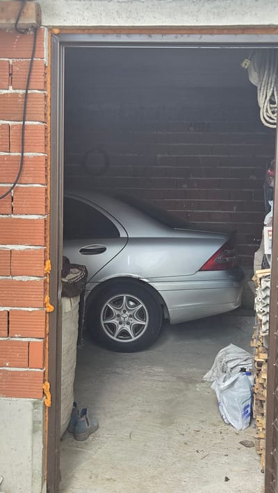 This media file shows the interior of a garage in Bania, Bulgaria. A silver sedan is parked inside, angled towards the camera. The car's rear is visible, with its taillight illuminated. A tire with a Mercedes-Benz rim is prominently displayed. The garage walls are made of brick, and the floor is concrete. Various objects are scattered around, including a coil of rope, a white plastic bag filled with items, and what appears to be a pair of shoes. The scene suggests a storage space or a place where a vehicle is being kept. There are no people visible in the image, and the overall impression is one of disarray or a cluttered environment. The lighting suggests it might be daytime, as there is ambient light filtering into the garage. The photograph seems to capture a moment of everyday life, highlighting the contents of a garage. No visible text is present in the image.