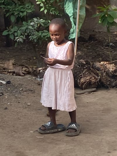 An outdoor, eye-level photograph captured in Udu, Nigeria, depicts a young Black child standing on bare earth. The child, with short dark hair, wears a light pink sleeveless top with a textured pattern and a matching plain light pink skirt. Oversized dark grey "adidas" brand slide sandals are on her feet. She is looking downwards with a focused expression, her mouth slightly ajar, as she manipulates a small, thin, light-colored object held between both hands at chest height. The background consists of green foliage on the left and right, with a notable green fabric draped vertically on the far right. The ground is irregular dirt, with scattered organic debris, including what appears to be a single brown boot lying on its side in the mid-left background. The scene is illuminated by natural daylight.