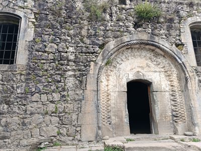 The image shows an ancient stone doorway with intricate carvings in the city of Khndzoresk, Armenia. The doorway is arched and features a rope-like border above a panel with a repeating geometric pattern. To the left of the doorway is a rough stone wall with a window barred by metal grates. The overall scene depicts an old, weathered structure, likely part of an ancient settlement or church, surrounded by natural elements like small plants growing in the crevices of the stonework. The ground in front of the doorway is made of stone steps. A timestamp "20 июля 2026 г. в 19:02" is superimposed on the lower part of the image, suggesting a capture date and time. The lighting indicates it is daytime.