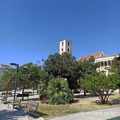 In a public square in Ruvo di Puglia, Italy, on July 6, 2026, at 09:38, the scene is captured under a clear blue sky. The foreground features paved pathways and several weathered wooden park benches. To the left, a modern street lamp stands. A variety of trees and shrubs dot the square, with a prominent palm tree in the center and a dense green tree to its right. In the background, a historic bell tower, constructed of light-colored stone with arched openings, rises above the foliage. Adjacent to the tower, a building with a red-tiled roof and a covered balcony supported by columns is visible. A sliver of the moon is present in the sky.
