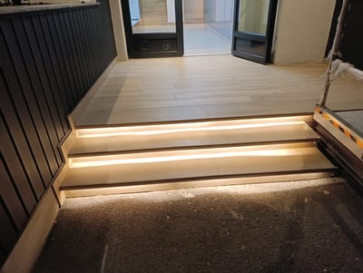 The image depicts a modern indoor staircase with integrated LED lighting. The stairs are constructed from light-colored wood, with each step featuring a recessed strip of warm white light. The wall to the left is clad in dark, vertical paneling. Beyond the staircase, a wide doorway leads to an area with light-colored wooden flooring and what appears to be glass doors. The foreground is a textured, dark surface, possibly gravel or a similar material. The scene is well-lit, highlighting the architectural features of the staircase and the transition to the adjoining space. This scene is located in Voluntari, Romania.
