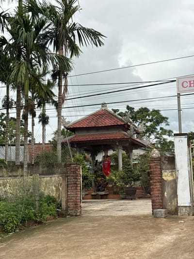 The image depicts a scene in Tân Lập Commune, Vietnam, dominated by lush greenery and traditional architectural elements. In the foreground, a dirt path leads towards an entrance framed by brick pillars and a weathered concrete wall. To the left, dense foliage and palm trees create a vibrant natural backdrop. A wire fence runs along the boundary.
The focal point of the scene is a traditional Vietnamese gazebo with a red tiled roof, adorned with intricate carvings. Inside the gazebo, a statue of a person in a red robe stands prominently. Various potted plants and trees are arranged around the gazebo, adding to the tranquil atmosphere.
Further back, more palm trees and a dense canopy of green trees are visible, suggesting a humid, tropical environment. Overhead, power lines crisscross the sky, a common sight in Vietnamese communities. A sign with pink lettering, partially visible on the right, indicates a commercial establishment. The sky is overcast, hinting at cloudy or potentially rainy weather. The overall impression is one of a serene, culturally rich location blending natural beauty with traditional architecture.