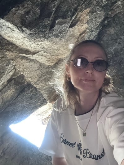 The image shows a young woman with blonde hair and sunglasses taking a selfie. She is wearing a white t-shirt with dark lettering that reads "Cornell Brand Est. 2014". She has a delicate silver chain with a small pendant around her neck. The woman is lying down in what appears to be a rocky environment, possibly a cave or crevice, as evidenced by the rough, textured stone walls surrounding her. Sunlight is streaming in from an opening, creating a bright, illuminated area in the lower right portion of the image. The lighting suggests it is daytime, and the overall mood seems relaxed and adventurous. The scene is set in Dom Otdykha Zolotoy Plyazh, Russia.