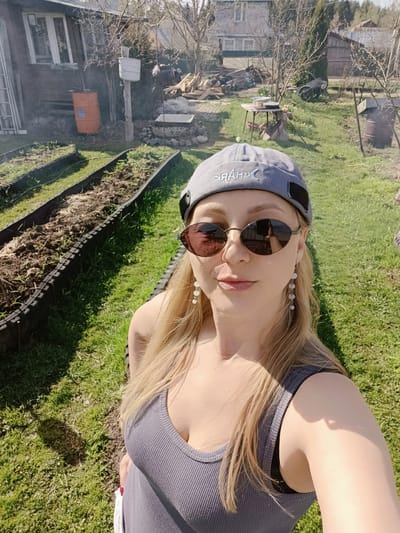 A woman takes a selfie outdoors. She is wearing sunglasses, a gray tank top, a gray cap with the word "SHARK" printed in reverse, and dangling earrings. She is standing on grass near garden beds and a house. In the background, there is a collection of firewood, other outbuildings, and trees. The image appears to be taken in Вичелово, Russia.