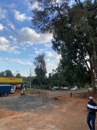 This image depicts a daytime street scene in Kianyaga, Kenya, characterized by a mix of commercial activity, construction, and natural vegetation under a sky with scattered cumulus clouds.
In the immediate foreground, a reddish-brown dirt ground occupies the lower portion of the frame. To the lower right, a man wearing a black and white long-sleeved shirt, dark blue jeans, and a white baseball cap is partially visible, facing left, holding a small red object in his right hand. To his left, a large mound of grey gravel is present, accompanied by a smaller pile of reddish-brown soil. A skeletal, angular metal frame, possibly for a structure or vehicle, lies on the dirt between the gravel and the paved road.
In the midground, a paved road extends horizontally across the scene. On the left side of the road, a yellow commercial building with red signage, partially obscured, indicates a shop or business. A blue box truck is parked directly in front of this building, with a white van or pickup truck parked behind it. A person wearing a high-visibility yellow-green vest is operating a motorcycle on a dirt patch adjacent to the commercial building, moving towards the right. Several other individuals are visible further down the road, including adults walking and children near the metal frame. Multiple vehicles are parked on the dirt shoulder and pavement on the right side of the road, including a red car with its boot open and several white SUVs or sedans. One adult male, dressed in a red shirt and dark pants, walks rightward on the dirt shoulder of the road.
The background is dominated by dense, tall green trees, some appearing mature with thick trunks and extensive canopies that frame the right side of the image and extend towards the top. Utility poles with power lines are visible against the sky on the left side. The distant landscape reveals additional trees and a slightly elevated terrain.