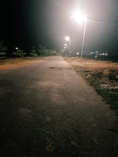 The image depicts a paved road at night, likely in a park or recreational area. The road stretches into the distance, lit by a series of streetlights. To the left of the road are trees and grass, while the right side shows dirt and grass. In the far distance, buildings can be seen under the glow of the streetlights. The road is cracked and shows signs of wear. The image is dark, with the streetlights providing the primary source of illumination. The sky is black and obscures any specific location in Rafin Pa, Nigeria. There are no people or vehicles visible in the frame.