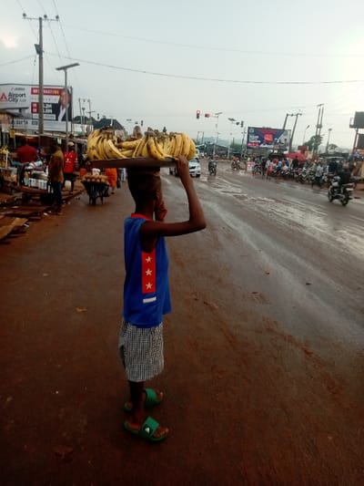 The image captures a candid moment of a young boy engaged in street vending, set against the backdrop of a bustling roadside scene.

In the foreground, slightly to the left of the center, a young boy stands facing away from the camera, looking towards the right side of the frame. He carries a large, flat tray piled high with ripe yellow bananas on his head. His left hand is raised, possibly steadying the tray or scratching his ear. He is wearing a blue tank top with a distinctive design on the right side: a vertical red stripe with three white stars stacked on it. Below this, he wears black and white checkered shorts and bright green slide sandals.

The ground where the boy stands and the road ahead appear to be reddish-brown, possibly a dirt or unpaved road, and show signs of being wet or muddy, with reflective patches suggesting puddles or recent rain.

To the left of the boy, further back, is the side of the road with several structures that appear to be market stalls or small shops. There are several adults visible, some standing near these stalls, and one person is pushing a handcart. Overhead, utility poles with numerous wires crisscross the sky. Two large advertising billboards are prominent on this side. One billboard, partially obscured, clearly displays the text "Airport City" and features a photograph of a man.

To the right of the boy, the road stretches into the distance, appearing busy with traffic. Several motorcycles are visible, some parked along the side, others in motion. At least one car can be seen. Further down the road, a traffic light shows a red signal. Another large billboard is visible further down the street on the right, with some text partially legible, including what appears to be "ABUJA TOGETHER". The sky above is overcast, suggesting either an early morning or late afternoon setting, with diffused light illuminating the scene. The overall impression is one of everyday activity in a vibrant, urban or semi-urban environment.