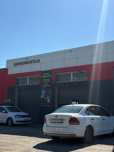 A white Volkswagen sedan is parked in front of a building with a sign that reads "Шиномонтаж" (Tire Fitting) in Russian. The building has a red and gray facade with several garage doors and windows. Another white car is parked to the left of the building. The scene appears to be a tire shop or car service center in Yagol, Russia, under a clear blue sky.