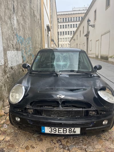 A black Mini Cooper, model generation R50/R52/R53, is parked on a cobblestone street. The vehicle displays significant front-end damage: its windshield is severely shattered with a prominent central impact point forming a spiderweb crack pattern; the hood is visibly dented; the front bumper is missing its upper grille, exposing the radiator; and the right (passenger-side) headlight is dislodged and angled downward, while the left (driver's-side) headlight is intact but heavily clouded. A Portuguese license plate, "P 39 EB 84," is affixed to the front bumper. The car's surface appears damp with water droplets.

To the immediate left of the vehicle is a tall, aged concrete wall with faded blue graffiti and a rectangular white patch. To the right, a narrow, cobblestone lane is bordered by a row of historic, light-colored buildings with multiple windows and an ornate black street lamp mounted on one facade. In the background, a large, multi-story institutional-style building with numerous small, rectangular windows dominates the skyline under an overcast sky. The scene is located on a narrow street in the city of Coimbra, Portugal, consistent with the architectural style and urban layout of its historic university district. No individuals are present.