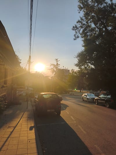 This is a photograph of a street scene in Harmanli, Bulgaria, during sunset. The sun is low on the horizon, casting long shadows and a warm, golden light across the scene. On the left, a building with a weathered facade is visible, its windows reflecting the sunlight. A sidewalk with tiled paving runs along the building, leading towards the foreground.
Several cars are parked on the right side of the street. In the foreground, a dark-colored SUV is parked, with its spare tire mounted on the rear. Further down the street, a silver hatchback and a black sedan are parked. Utility poles with numerous overhead wires are also present, contributing to the urban atmosphere.
In the background, trees and more distant buildings are silhouetted against the bright sky. The overall impression is one of a quiet, late afternoon or early evening in a residential or mixed-use urban area. The image is captured with a slight tilt, adding a dynamic quality to the composition.