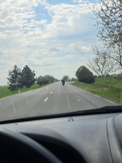 View from inside a vehicle traveling on a paved road outside Gorno Aleksandrovo, Bulgaria. The road has a white centerline and green fields on either side, lined with trees. A motorcycle is approaching in the distance. The sky is partly cloudy. The interior of the vehicle is visible on the right.