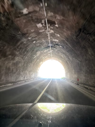 The image is a point-of-view shot from inside a vehicle traveling through a tunnel. The tunnel walls are rough, rocky, and uneven, with some patches of lighter colored material. There are lights running along the ceiling of the tunnel. The road ahead is asphalt and has a white line marking the center. The end of the tunnel is brightly lit, suggesting an exit to daylight. The foreground shows the hood and windshield of the vehicle, with a reflection of the outside environment visible on the hood. The scene is dark and appears to be in a mountainous or rocky area. The image does not contain any specific landmarks or clues to identify its location within Mizur, Russia.
