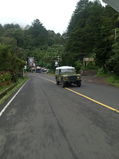 The scene depicts a two-lane asphalt road in Deothang, Bhutan, winding through a densely vegetated, mountainous landscape under an overcast sky. An olive green, Land Rover-style vehicle with a white hardtop is in motion, positioned on the right side of the road and traveling away from the viewer. The road features a solid yellow center line and a solid white edge line on the left. Tire marks are visible on the asphalt surface.
To the left, beyond a slight curve in the road, a cluster of small, single-story structures, possibly shops, are visible, fronted by utility poles and multiple dark banners with white markings. Dense green trees and foliage cover the steep hillsides on both sides of the road, extending upwards. On the right, a dilapidated building with an exposed frame and a partially legible sign (appearing to contain "INHERITANCE") stands amidst the trees, accompanied by a low, graffitied barrier. No human subjects are present in the image.