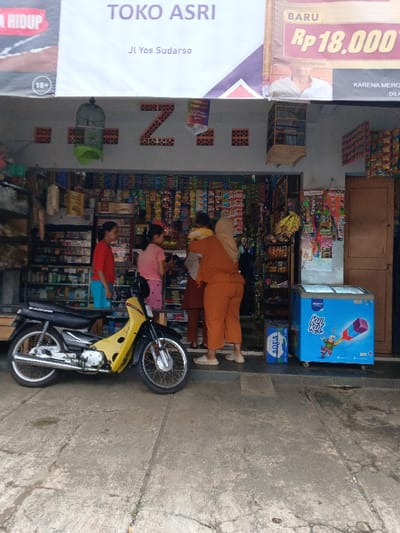 This image captures the facade and interior of "Toko Asri," a small convenience shop located on Jl Yos Sudarso in Ciamis, Indonesia. The scene appears to be daytime, with bright, diffused natural light illuminating the area.

In front of the shop, a yellow and black motorcycle is parked on the concrete ground. The shop's entrance is open, revealing shelves densely packed with various packaged goods, snacks, and household items. Four individuals are visible within or near the entrance. On the left, a woman in a red shirt and blue shorts stands inside, looking towards the shop's interior. Next to her, a girl in a pink shirt also looks inward. With their backs to the camera, two other individuals are positioned further inside the shop: one in an orange outfit, and another in a brown outfit and a light-colored hijab, both seemingly browsing or making a purchase.

To the right of the entrance, a stack of AQUA bottled water is visible, next to a blue ice cream freezer with "Kul Kul" branding. The shop's facade features a decorative "Z" shape made from bricks and two hanging birdcages. Above the entrance, a large banner displays "TOKO ASRI" and "Jl Yos Sudarso." Other visible text includes "BARU Rp 18,000" and partial words like "HIDUP," "18+," "KARENA MERCO," and "DILA" on various banners and signs, indicating advertisements for products or services.