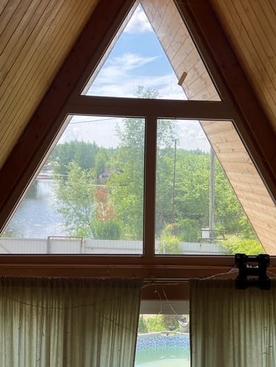 The image shows a view through a large, triangular window of a house. The window is divided into multiple panes and offers a view of a lake, trees, and a fence. The interior of the house is visible in the foreground, with wooden paneling on the ceiling and walls. There are light-colored curtains hanging below the window, and a small section of a swimming pool is visible through a lower opening. Outside, a body of water, likely a lake, is visible on the left, with a faint reflection of houses and trees on its surface. A white fence runs along the edge of the property, separating it from the water. Lush green trees and vegetation surround the lake, with some houses visible in the distance. A few electrical poles are also present in the landscape. The sky is partly cloudy. The scene appears to be set in a rural or semi-rural location. Specifically, the location is identified as Vasilyevo, Russia, suggesting a view from a house in that city.