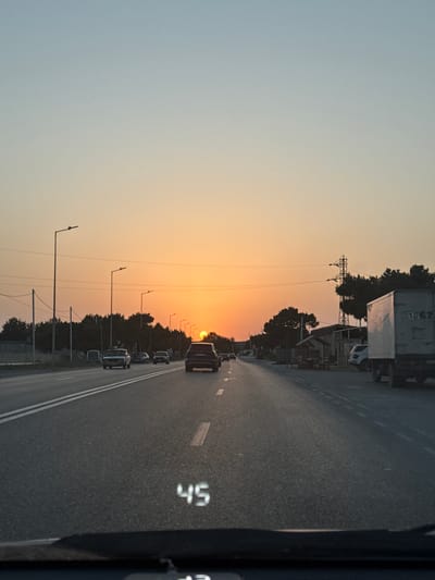 The image depicts a road scene at dusk, likely in the city of Mstaga, Azerbaijan, given the context. The sun is low on the horizon, casting a warm orange glow across the sky and reflecting on the asphalt road. Several vehicles are visible on the road, including cars and a white truck. The road is paved and has white lane markings. On the left side of the road, there are trees and utility poles with power lines. On the right side, there are some small structures, more trees, and a large electrical pylon. A digital display on the windshield of the vehicle from which the photo is taken shows "45", indicating the speed. The overall scene suggests a typical evening commute or travel on a highway.