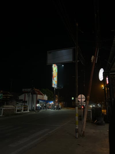 The image depicts a nighttime street scene in Crewek, Indonesia. A road with a pedestrian crossing is visible in the foreground, illuminated by streetlights and lights from nearby establishments. On the left side of the road, a building with the sign "SMK PGRI KUWU" is present. Further down the street, there are small shops or food stalls with some individuals present. A large billboard with a colorful advertisement is illuminated, and a blank billboard is positioned above it. Electrical poles with numerous wires are prominent features throughout the scene. The overall environment is dark, typical of a rural or semi-urban area at night.