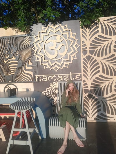 A young woman with long blonde hair sits barefoot on a slatted chair. She is wearing a green hooded dress and is holding a phone to her ear, looking towards the camera. Behind her is a large, decorative wooden wall featuring an Om symbol, the word "Namaste" in stylized lettering, and intricate leaf-like patterns. To her left, a light blue table and a grey slatted stool are visible. The scene is outdoors, with trees and blue sky visible at the top of the frame. The lighting suggests it is daytime, with shadows cast by the trees and the wall. The setting appears to be a public space, possibly a park or an outdoor event area in Голубинское сельское поселение, Ukraine. The woman's expression is neutral.