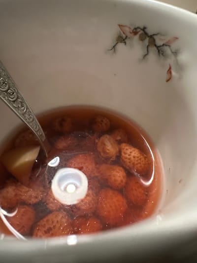 A close-up, slightly blurry photograph shows a white ceramic teacup filled with a reddish liquid, likely tea or a compote, and several small, whole strawberries. A portion of a silver spoon with an ornate handle is visible in the upper left quadrant of the image, resting inside the cup. The teacup itself has a delicate, faded floral pattern on its upper rim. The lighting suggests an indoor setting, possibly a kitchen or dining area. There are no people visible in the image. The location of this scene in the city of Biysk, Russia, is not ascertainable from the visual information provided in the image.