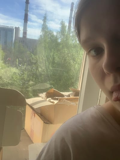 The image appears to be a selfie taken indoors, looking out a window. The individual's face is partially visible in the upper portion of the frame, with one eye prominently displayed. A portion of their tongue is also visible. The background is dominated by an outdoor scene, visible through the window. The scene includes foliage, likely trees, and an industrial structure with a tall chimney. The chimney has alternating red and white sections. The building is labeled "КОМПЛЕКС H49," indicating the location is likely in or near an industrial complex. Based on this label and the appearance of the chimney, the industrial complex is located in Novoe Devyatkino, Russia. To the right side of the window, inside the room, there are cardboard boxes. One box appears to be partially open and torn, with remnants of packaging materials visible. Part of a light colored chair is also visible in the lower part of the frame. The lighting suggests it is daytime.