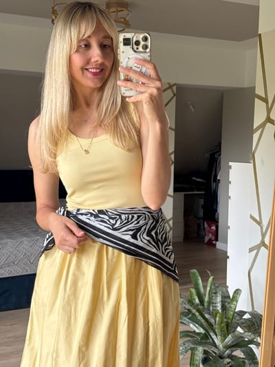 A woman with blonde hair and bangs, wearing a yellow tank top and a yellow skirt with a zebra-print scarf tied around her waist, takes a mirror selfie in a bedroom. The room appears to be in a residential setting, possibly an apartment in Carnikava, Latvia. In the background, a bed with a grey patterned comforter is visible, along with a closet with clothes. A potted plant sits to the right of the frame, and a mirror with a wooden frame is positioned prominently. The woman is holding a smartphone, which is decorated with stickers. The lighting suggests it is daytime.
