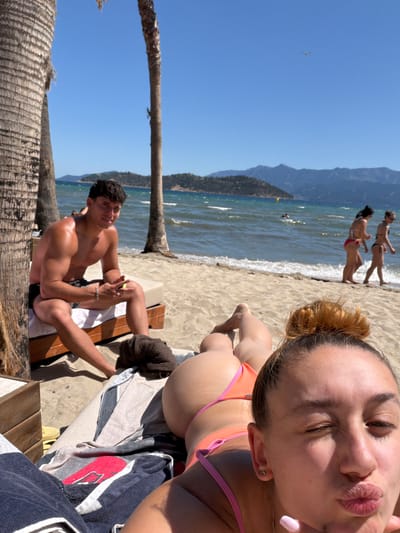 In Keramoti, Greece, a sunny day at the beach unfolds. A man, shirtless and muscular, sits on a lounge chair, holding a mobile phone and a small yellow object. He wears black swim trunks and a silver bracelet. Two palm trees frame the scene, with their rough bark contrasting against the bright blue sky. In the background, the choppy, turquoise water of the sea is visible, with gentle waves lapping the sandy shore. Several people are in the water, including one person swimming. Further out, a range of hazy, blue-green mountains forms the horizon. In the foreground, a woman lies on a towel, her back to the camera, wearing an orange and pink bikini. Her head is turned, and she is making a kissing face towards the camera, winking. Her hair is pulled up in a bun, and she has light pink nail polish on her fingernails. A dark towel rests on the sand near the man. The overall atmosphere is one of leisure and relaxation on a warm, summer day.