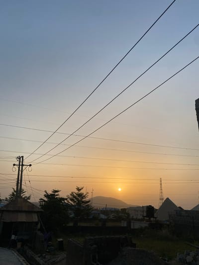 A low-angle, rotated landscape captures a hazy, golden sunrise or sunset behind a distant mountain. Multiple power lines stretch across the frame, converging towards the bottom right corner, where a utility pole stands near a house. Green trees and buildings are vaguely visible, and a transmission tower sits far off in the background to the right of the sun. The color scheme is dominated by yellow, orange, and gray, creating a muted and hazy atmosphere.