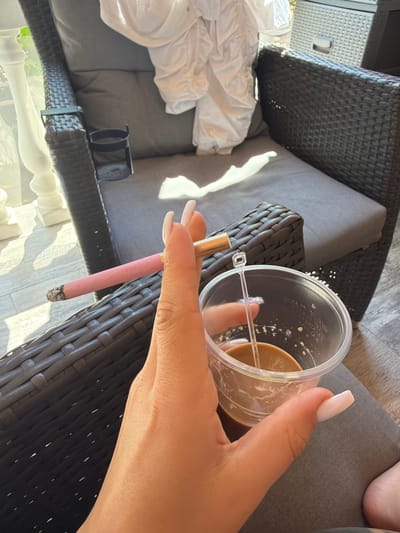 A person's hand holds a pink cigarette and a clear plastic cup of coffee with a stirrer in it. The person is sitting in a dark wicker chair with gray cushions. In the background, there is a white chair with white cloth draped over it, and a white railing. The scene appears to be outdoors on a sunny day. The location is in Novi Iskar, Bulgaria.