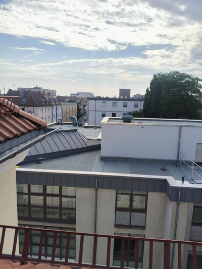 This is a vertical, eye-level, medium shot of a cityscape taken from a rooftop. The sky is a pale blue with scattered, wispy white clouds. The sun is shining, indicating it is daytime, likely late morning or early afternoon, judging by the brightness.
The foreground is dominated by the rooftop itself, with various buildings and structures. To the left, a reddish-brown tiled roof with a metal gutter runs diagonally across the frame. Below this, a light-colored building with a textured facade is visible, featuring a row of windows with red crosses and text that reads "Bayerisches Rotes Kreuz". Further into the frame, more rooftops are visible, covered in dark grey, flat roofing material. These include a building with a light grey, angular roof structure and several ventilation units. On the right, a larger, modern white building with a flat roof has pipes and railings on its exterior.
In the background, a dense cluster of multi-story buildings can be seen, with a variety of architectural styles and colors, ranging from light yellow to white and grey. A large, mature green tree stands prominently on the right side of the background, its leaves lush and full. In the far distance, taller structures, possibly commercial buildings or towers, are silhouetted against the sky.
There are no people visible in the image. The scene appears to be a quiet urban environment, with a focus on the architecture and the sky. The presence of the "Bayerisches Rotes Kreuz" text suggests a location associated with the German Red Cross, and the Nuremberg context places the scene within that specific German city. The overall mood is serene and observational, capturing a typical urban view under a partly cloudy sky.