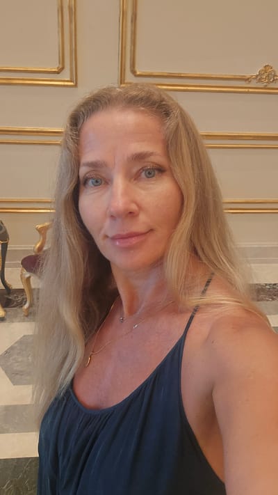 A middle-aged Caucasian woman with long blonde hair and blue eyes is pictured in a selfie. She is wearing a dark blue spaghetti strap top and a delicate gold necklace with a small pendant. The background suggests an indoor setting with ornate gold trim on cream-colored walls and a patterned tiled floor. There is a hint of a chair with red upholstery and gold accents to her left. The lighting is soft, and the woman's expression is neutral. The location is the city of Agios Epiktitos, Northern Cyprus / Cyprus.