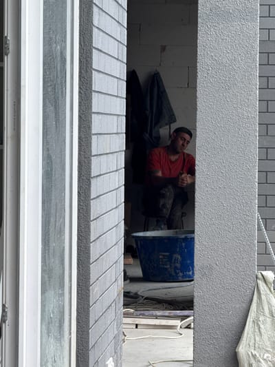 A man wearing a red t-shirt and dark pants sits on a stool inside a building under construction. He has a cap on his head and is looking down at his hands, which are clasped in front of him. A large blue bucket is on the floor in front of him, and construction materials are visible in the background. The scene appears to be a construction site in Yalta, Ukraine.