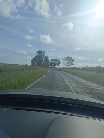 This is a forward-facing, point-of-view shot from inside a vehicle, looking out at a paved rural road on a bright, partly cloudy day. The road curves to the left and is bordered by grassy verges and fields. Several mature trees are visible on either side of the road, with the sun, partially obscured by clouds, shining brightly in the upper right portion of the frame, creating lens flare. The foreground shows the dashboard of the vehicle. The scene is consistent with a drive through the countryside of Huntingdonshire, United Kingdom, given the typical landscape features and atmospheric conditions for the region.