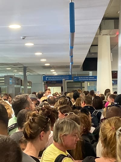 This image depicts a crowded indoor environment, likely an airport terminal, with numerous people standing in a queue. Several signs are visible in the background, including one that reads "Medunarodni odlaci International Departures." The lighting appears to be artificial, coming from ceiling fixtures. The scene is densely populated, with individuals facing in various directions, suggesting a waiting area or a security checkpoint. Some people are looking at their phones, while others appear to be conversing or looking towards the front of the line. The overall atmosphere is one of congestion and waiting. The location is identified as Mrčevac, Montenegro, indicating this is likely part of Tivat Airport.