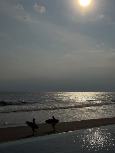 A backlit coastal scene is depicted in Belalang, Indonesia. The bright, intense sun, positioned in the upper right, illuminates a hazy, pale blue-grey sky with scattered clouds. The ocean to the right exhibits a broad, sparkling path of sunlight reflecting on its surface, extending to the horizon. Small waves break along the dark sand shoreline. Two individuals, appearing as dark silhouettes, traverse the beach from left to right, each carrying a surfboard. The surfboard carried by the person on the left is visibly lighter in tone than the dark board held by the person on the right. In the immediate foreground, a body of water, likely an infinity pool, glistens with numerous bright reflections from the overhead sun.