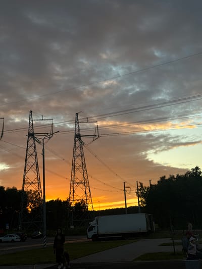 This media file captures a sunset scene in Bobrovo, Russia. The sky is a dramatic canvas of orange and yellow hues, contrasting with dark, ominous clouds. Towering power lines and their supporting pylons dominate the foreground, silhouetted against the vibrant sky. In the background, a line of dark trees separates the sky from a road or pathway.

On the right side of the image, a paved path runs alongside a grassy area. A white truck is parked near the path, with its back doors open. Further down the path, a person is visible, appearing to be walking or standing. A smaller white vehicle is parked on the road.

The time of day is clearly sunset, indicated by the intense colors in the sky. The weather appears to be mixed, with the stormy clouds suggesting recent or approaching rain, yet the sunset is still brilliant. The presence of power lines suggests an area with infrastructure, possibly a suburban or rural setting with utility access. The overall mood is one of natural beauty juxtaposed with man-made structures. There is no visible text in the image.