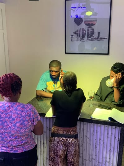 An indoor scene depicts four individuals gathered around a dark-speckled counter. On the left, a female with reddish-purple braided hair in a bun wears a floral top, her back to the camera. To her right, a second female with dark braided hair, wearing a black top and leopard-print pants, leans forward, reviewing papers on the counter. Opposite them, two males are positioned at the counter. The male in the center, wearing a teal t-shirt, holds a dark smartphone to his right ear, engaged in a conversation, with another phone resting on the counter. The male on the right, wearing a dark long-sleeved shirt and sunglasses on his head, holds a small white portable fan to his face. A wine glass containing red liquid, an open notebook, and a black smartphone are arranged on the counter in front of him. The background wall is light-colored and features a large framed picture depicting various glasses and dark fruits. The lower portion of the counter's front is paneled with a light-colored, marble-patterned material, illuminated by a subtle purple light. A yellowish-green light source illuminates the wall area directly behind the two males. The specific location within Moba, Nigeria, is not evident from the image.