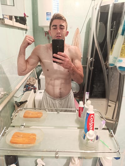 A shirtless man with defined abs is taking a mirror selfie in a bathroom. He is flexing his bicep on the left side of the image and holding his phone in his right hand to capture the reflection. He is wearing light-colored shorts.
In the background, a shower stall is visible, along with a washing machine and a water heater. A radiator with some clothing draped over it is also present. A shelf in front of the mirror holds two pieces of what appears to be raw salmon on clear plates, a toothbrush, and tubes of Colgate toothpaste. Another shelf below has a black bottle and a white pump dispenser.
The bathroom appears to be tiled with light green tiles. The lighting suggests it is indoors. There is text on the toothpaste tubes, with "Colgate" being clearly legible. The water heater also has some text and diagrams on it.