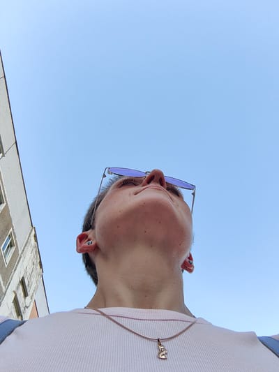 A low-angle, waist-up shot shows a person looking up at a clear blue sky. The person has short brown hair and is wearing purple-tinted sunglasses, a pale pink ribbed t-shirt, and a silver necklace with a cat-shaped pendant. They have two piercings in their left ear, each with a small blue stud. To the left of the person is the side of a weathered, multi-story building with visible windows. The scene suggests it is daytime, with bright overhead lighting and a clear sky, indicating fair weather. The setting appears to be an urban environment, possibly a street or courtyard, given the presence of the building. The person's upward gaze and the upward camera angle create a sense of contemplation or observation of the sky and their surroundings. No text is visible in the image.