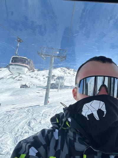A selfie image captured from inside a gondola lift, depicting a person in ski attire overlooking a snow-covered mountain landscape in Les Allues, France. The subject, visible from the forehead to the upper torso, occupies the right foreground. They wear large, dark-framed, mirrored ski goggles that obscure their eyes, a black neck gaiter featuring a white, stylized, geometric elephant logo, and a ski jacket with a camouflage-like pattern in grey, black, and white, accented with neon yellow. The interior view is through a transparent, scratched window.
Beyond the window, a bright, clear blue sky is visible above snow-covered mountains. To the left, another white gondola, prominently labeled "Les vallées" and partially "Méribel-Mottaret," hangs from a cable, moving across the frame. Further into the mid-ground, a tall grey ski lift pylon, marked "U5," stands on the snowy slope. The mountainous terrain is extensively covered in snow, revealing rocky outcrops and visible ski tracks. Additional ski infrastructure, including buildings and support structures, is discernible on the distant slopes. The scene is illuminated by bright sunlight, indicative of clear weather conditions.