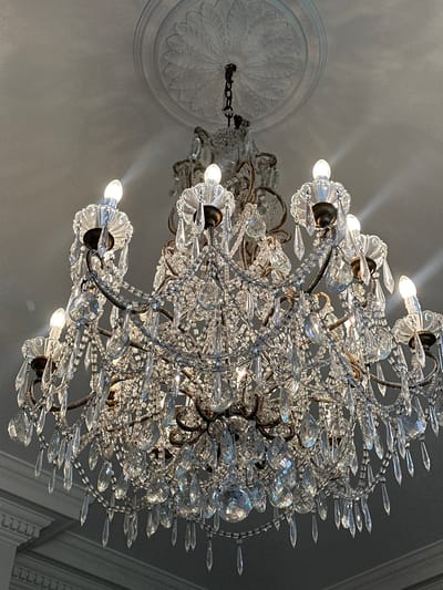 The image shows a large, ornate crystal chandelier hanging from a ceiling. The chandelier has multiple tiers and is adorned with numerous glass prisms and beads. Several light bulbs are visible, illuminating the crystals and creating a shimmering effect. The ceiling features a decorative plaster medallion around the point where the chandelier is attached. The overall impression is one of grandeur and elegance. The image does not provide any information about the city of Chichester, United Kingdom, or any specific location within it. There are no people or actions depicted in the image.