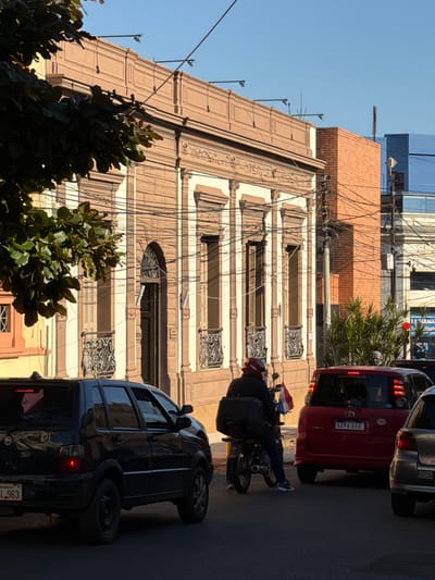 The image shows a street scene in Asuncion, Paraguay, with a focus on a building's facade and vehicles. The building appears to be old, with ornate details around its windows and a warm, tan-colored facade. A red car is parked on the right side of the image, and a dark-colored SUV is also visible. A person is riding a motorcycle, carrying what looks like a black bag on the back. The motorcycle has a flag attached to it. The sky is a clear blue, suggesting it's a sunny day. The presence of many overhead wires indicates a possibly older infrastructure. The time of day appears to be late afternoon due to the shadows cast. The license plate on the red car reads "AAPA 888". The license plate on the dark SUV is partially visible and ends with "L.963". The scene conveys a typical urban street view with daily life unfolding.